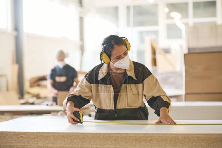 Téléchargez les photos : Female craftsperson in protective mask and headphones using tape measure to examine the length of wooden plank - en image libre de droit