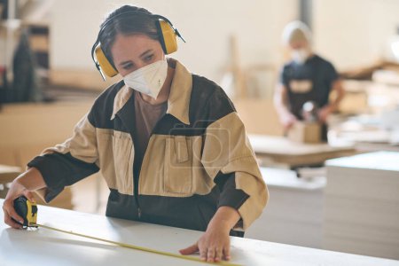 Foto de Young female worker in protective mask and headphones measuring the plank with tape measure during work at factory - Imagen libre de derechos