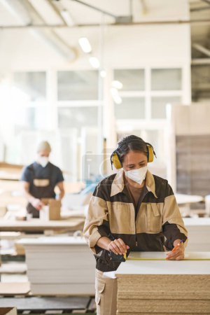 Foto de Trabajadora joven en máscara y auriculares midiendo tablón de madera antes de aserrar durante su trabajo en fábrica - Imagen libre de derechos