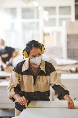 Foto de Carpintero femenino en máscara protectora y auriculares haciendo medidas de tablas de madera con regla en fábrica - Imagen libre de derechos