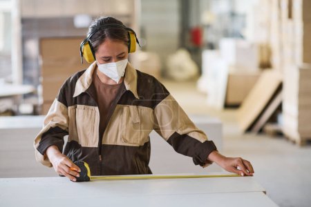 Téléchargez les photos : Jeune travailleuse masquée et uniforme effectuant des mesures avec ruban à mesurer pendant son travail à l'usine - en image libre de droit