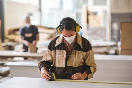 Foto de Young female worker in protective mask and headphones using tape measure to make measurements on wooden planks - Imagen libre de derechos