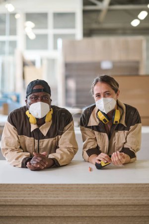 Foto de Retrato de dos colegas en máscaras protectoras mirando a la cámara mientras trabajan juntos en la fábrica de muebles - Imagen libre de derechos