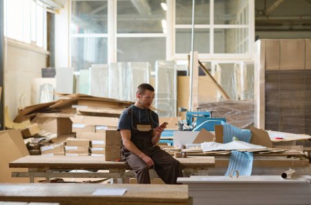 Téléchargez les photos : Ouvrier assis sur la table et utilisant son téléphone portable pendant sa pause après le travail dans un entrepôt - en image libre de droit