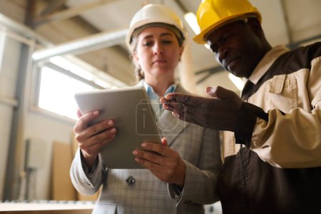 Photo for African foreman in hardhat pointing at tablet pc and talking to engineer during work at factory - Royalty Free Image
