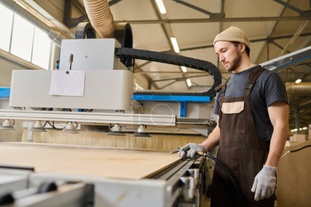 Téléchargez les photos : Jeune menuisier en tablier travaillant avec des planches de bois à la machine moderne à l'usine de meubles - en image libre de droit