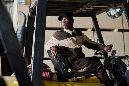 Photo for African male worker in uniform driving forklift and transporting freight in warehouse - Royalty Free Image
