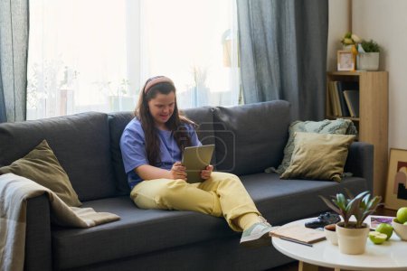 Photo for Happy girl with Down syndrome using tablet while relaxing on couch in living room at leisure and communicating in video chat - Royalty Free Image