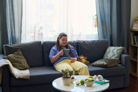 Photo for Young female with cup of tea or coffee relaxing o nsoft comfortable couch in living room at leisure while watching movie on tv - Royalty Free Image
