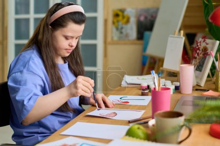 Foto de Chica con discapacidad mental pintura con acuarelas y pincel sobre papel mientras está sentado junto al escritorio delante de la computadora portátil en la sala de estar - Imagen libre de derechos