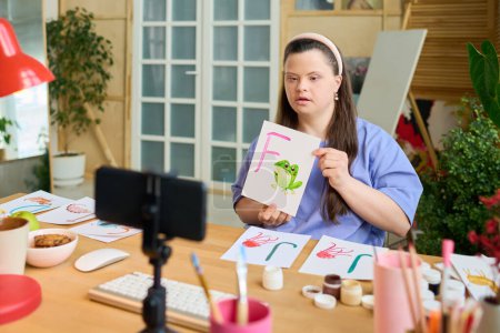 Foto de Mujer joven con síndrome de Down mostrando papel con rana verde pintada y letra F mientras se comunica con el público en línea - Imagen libre de derechos