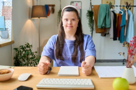 Photo for Happy young female with Down syndrome sitting by desk in front of computer ready for online lesson and looking at you - Royalty Free Image