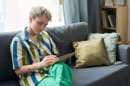 Foto de Joven mujer seria con discapacidad dibujando bocetos en bloc de notas mientras está sentado en la suave y cómoda soafa con cojines en la sala de estar - Imagen libre de derechos