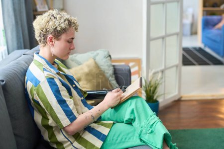 Foto de Joven amputada en ropa casual haciendo bocetos con lápiz en bloc de notas mientras está sentada en un sofá suave en el ambiente hogareño - Imagen libre de derechos