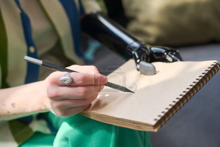 Téléchargez les photos : Main d'une jeune femme handicapée physique tenant un crayon sur une page blanche du bloc-notes assis sur un canapé et dessinant un croquis - en image libre de droit