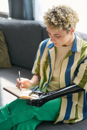 Téléchargez les photos : Jeune femme handicapée en tenue décontractée assise sur un canapé confortable dans le salon et dessin au crayon dans un bloc-notes - en image libre de droit