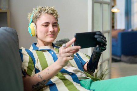 Foto de Joven mujer sonriente en ropa de casualwear mirando a la pantalla del teléfono inteligente en sus manos mientras se relaja en el sofá y se comunica en el chat de vídeo - Imagen libre de derechos
