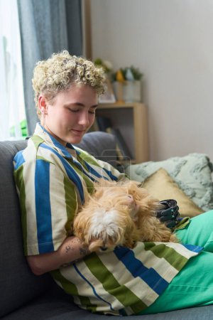 Téléchargez les photos : Fille heureuse avec handicap physique jouer avec chien de race mignon tout en se relaxant sur un canapé confortable dans le salon à loisir - en image libre de droit
