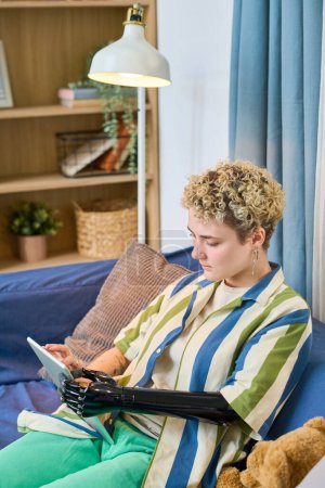 Téléchargez les photos : Jeune femme avec bras myoélectrique utilisant une tablette numérique pour la communication en ligne tout en se relaxant sur un canapé confortable dans le salon - en image libre de droit