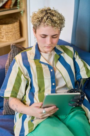 Téléchargez les photos : Jeune femme sérieuse avec prothèse de bras regardant l'écran de la tablette tout en étant assis sur un canapé confortable dans le salon et en travaillant dans le filet - en image libre de droit