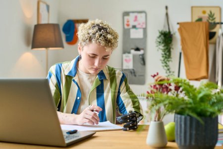 Téléchargez les photos : Jeune femme d'affaires moderne ou employée avec un bras partiel regardant à travers les documents financiers tout en travaillant au bureau à domicile - en image libre de droit