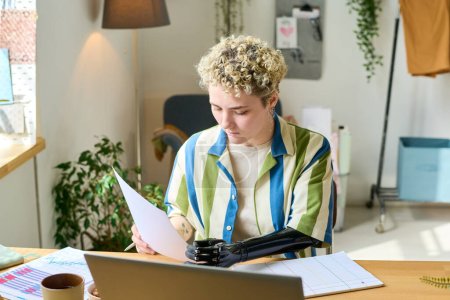 Junge Ökonomin mit Armprothese arbeitet mit Finanzdokumenten, während sie zu Hause am Arbeitsplatz vor dem Laptop sitzt