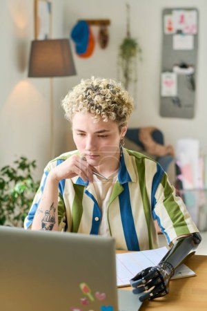 Téléchargez les photos : Jeune femme sérieuse avec bras myoélectrique regardant écran d'ordinateur portable tout en travaillant sur un nouveau projet d'entreprise par bureau dans le bureau à la maison - en image libre de droit