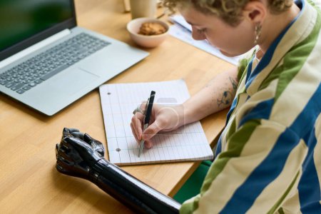 Téléchargez les photos : Jeune femme d'affaires avec bras partiel prendre des notes ou écrire un plan de travail pour la semaine tout en étant assis par bureau devant un ordinateur portable - en image libre de droit