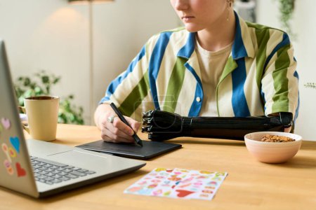 Foto de Joven diseñador gráfico con lápiz óptico y tableta gráfica retocando imágenes o dibujo mientras está sentado junto al escritorio frente a la computadora portátil en la oficina en casa - Imagen libre de derechos