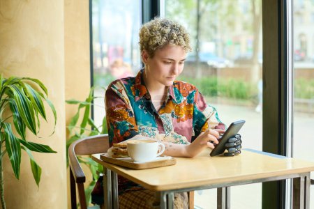 Foto de Mujer joven con discapacidad física desplazándose en el teléfono inteligente mientras está sentada junto a la mesa en la cafetería y tomando café con sándwiches - Imagen libre de derechos