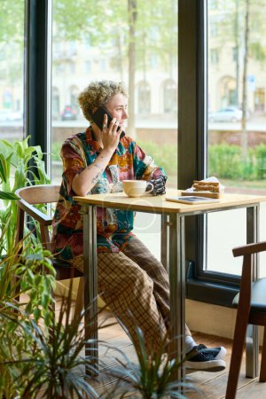 Téléchargez les photos : Jeune femme avec bras partiel parlant sur téléphone portable à table dans un café et regardant par une grande fenêtre tout en se relaxant à loisir - en image libre de droit