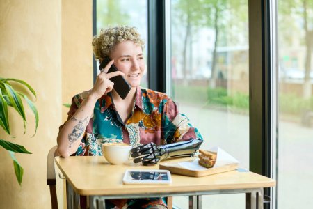 Téléchargez les photos : Joyeux jeune femme avec bras partiel parlant sur un téléphone portable assis à table dans un café, regardant par la fenêtre et prenant un café - en image libre de droit