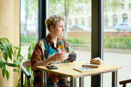 Téléchargez les photos : Jeune femme sereine avec une déficience physique regardant par une grande fenêtre alors qu'elle était assise à table dans un café et prenant un café avec collation - en image libre de droit