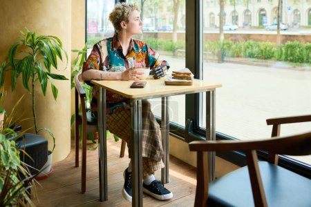 Téléchargez les photos : Jeune femme en tenue décontractée regardant par la fenêtre tout en étant assis à table dans un café confortable et prendre un café avec collation pour le petit déjeuner - en image libre de droit
