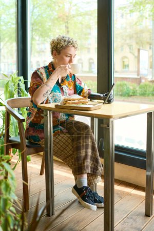 Foto de Joven hembra descansada con brazo mioeléctrico usando smartphone y tomando café con sándwich para el desayuno mientras está sentada a la mesa en la cafetería - Imagen libre de derechos