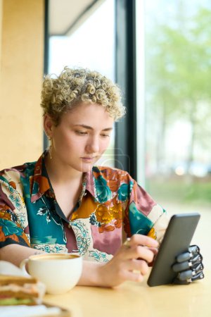 Téléchargez les photos : Jeune femme avec bras partiel défilant à travers des vidéos en ligne dans son smartphone tout en se relaxant dans un café confortable et en prenant un café - en image libre de droit