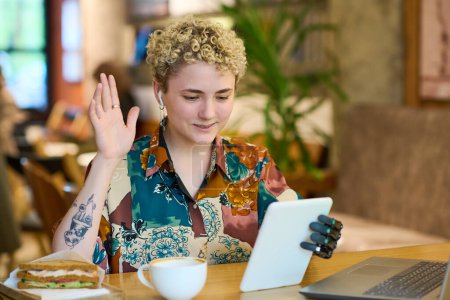 Téléchargez les photos : Jeune femme souriante avec handicap agitant la main et dire bonjour à quelqu'un sur l'écran de la tablette tout en communiquant dans le chat vidéo dans le café - en image libre de droit