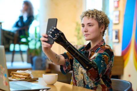 Foto de Mujer joven con brazo robótico mirando a la cámara del teléfono inteligente mientras se hace selfie o se comunica en el chat de vídeo durante el desayuno - Imagen libre de derechos