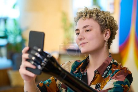 Foto de Chica rubia feliz con brazo mioeléctrico haciendo selfie mientras descansa y merienda en la acogedora cafetería y mirando en la cámara del teléfono inteligente - Imagen libre de derechos