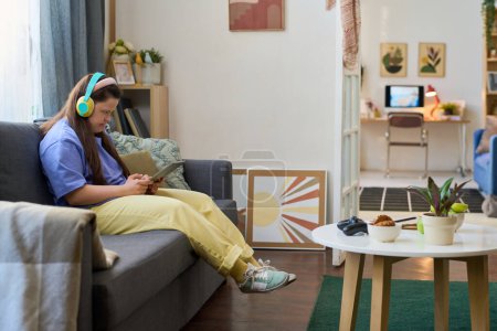 Téléchargez les photos : Fille avec le syndrome de Down dans les écouteurs regardant l'écran de la tablette pendant la communication dans le chat vidéo tout en étant assis sur le canapé à la maison - en image libre de droit