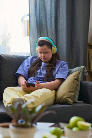 Photo for Young female with Down syndrome in headphones looking at smartphone screen while sitting on comfortable couch at home - Royalty Free Image