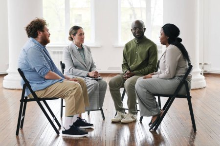 Foto de Grupo de personas interracial escuchando a una joven mujer afroamericana paciente de psicoterapeuta sentada en sillas en círculo - Imagen libre de derechos