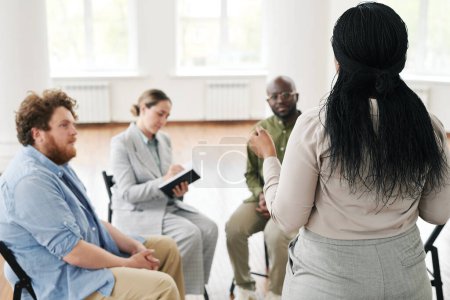 Téléchargez les photos : Dos de la femme noire décrivant ses problèmes tout en se tenant devant un groupe de patients assistant à des cours de séances psychologiques - en image libre de droit