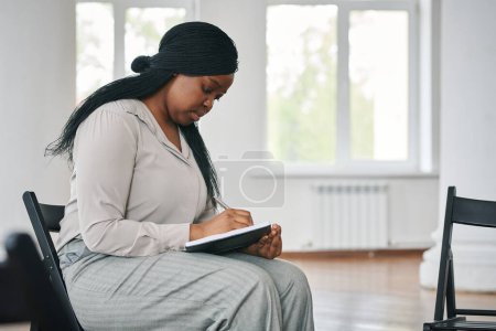 Foto de Joven mujer negra seria haciendo notas en el cuaderno antes o después de la sesión de psicoterapia mientras está sentado en la sala de conferencias espaciosa - Imagen libre de derechos