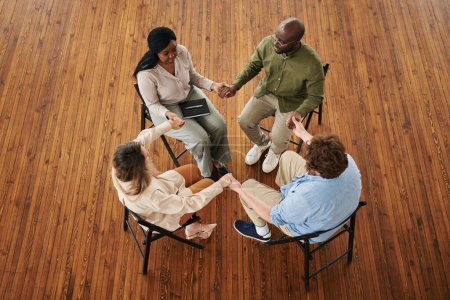 Photo for Group of young interracial people holding by hands while sitting on chairs during session, discussing their problems and supporting each other - Royalty Free Image