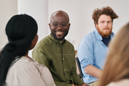 Foto de Feliz joven negro mirando a uno de los pacientes durante la sesión psicológica mientras se sienta entre otras personas y la escucha - Imagen libre de derechos