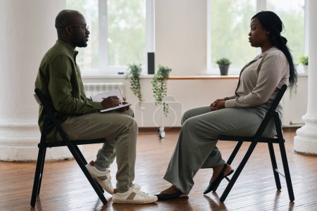 Foto de Vista lateral de un joven psicólogo afroamericano con un cuaderno consultando a una paciente sentada frente a él en la sesión - Imagen libre de derechos