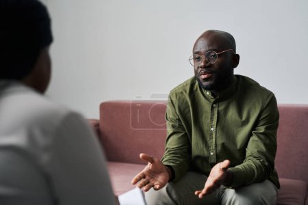 Foto de Joven hombre negro en ropa casual sentado en el sofá y explicando sus emociones y preocupaciones durante la charla con el psicoanalista - Imagen libre de derechos