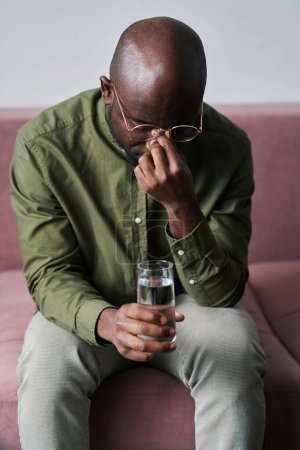 Foto de Joven hombre afroamericano preocupado con un vaso de agua sentado en el sofá y tocando el puente nasal mientras asistía al curso psicológico - Imagen libre de derechos