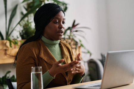 Téléchargez les photos : Jeune psychanalyste afro-américain confiant assis par bureau devant un ordinateur portable et parlant au patient pendant une session psychologique en ligne - en image libre de droit